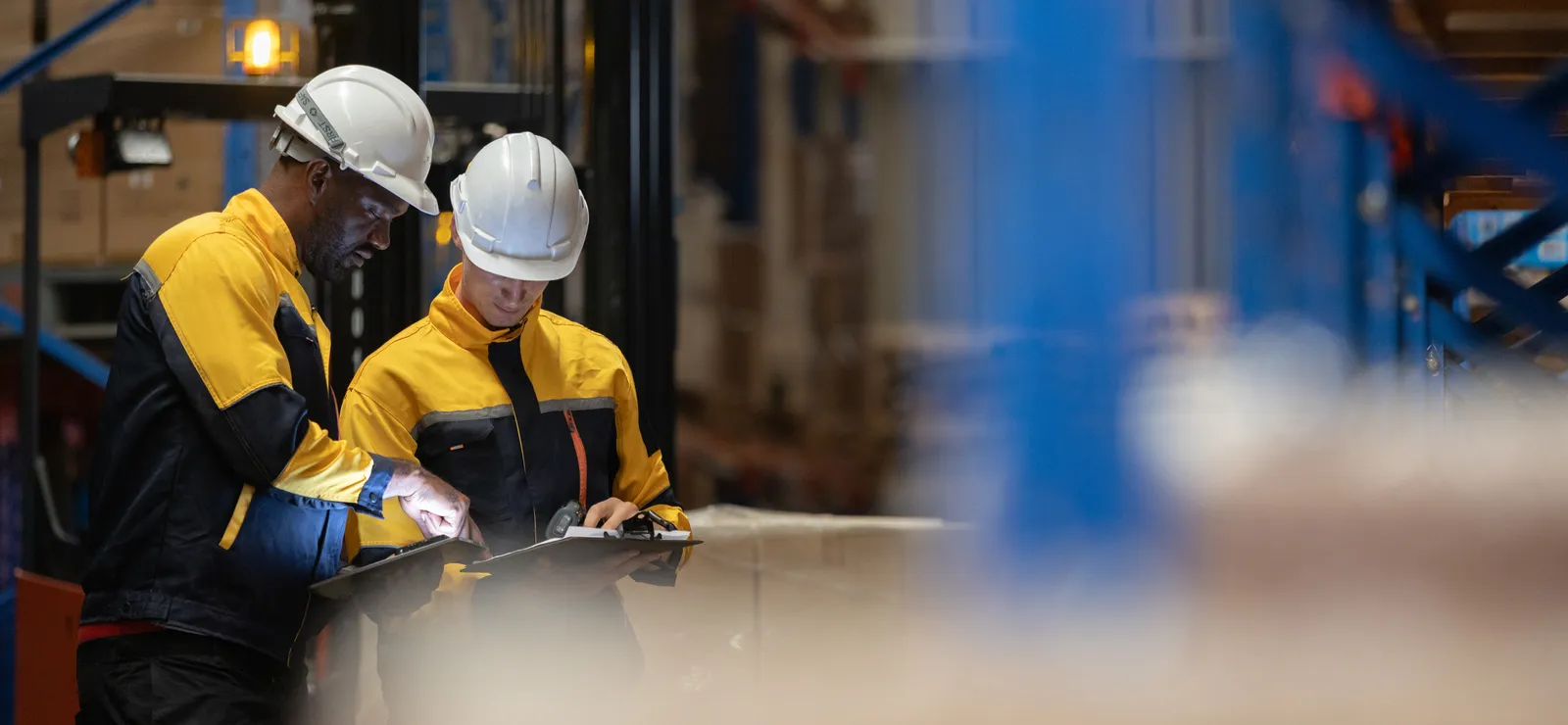 Two warehouse workers in hard hats reviewing order sheets on clipboards